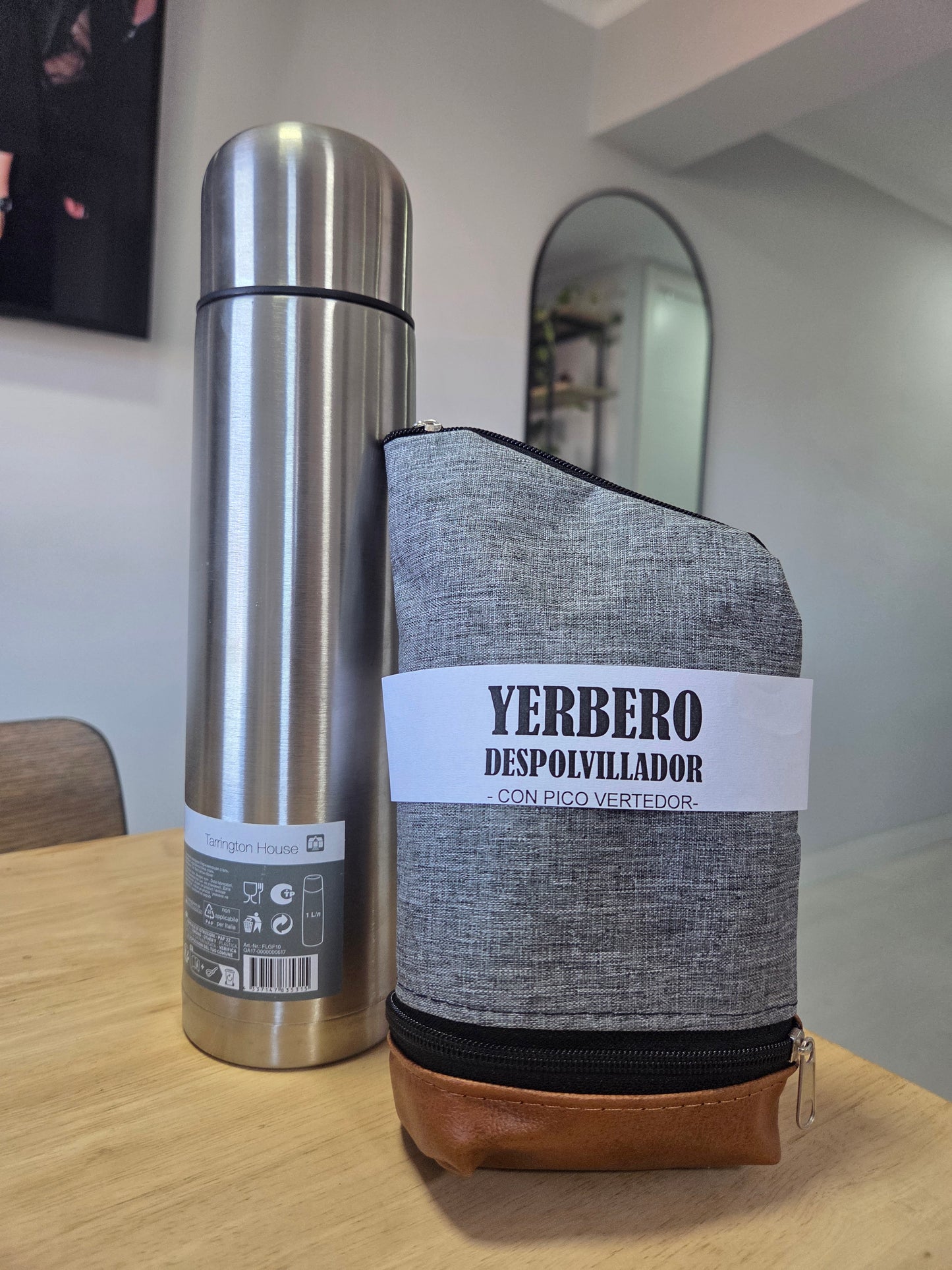 Yerbero despolvillador de tela gris con pico vertedor junto a un termo de acero inoxidable sobre una mesa.